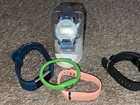 Five Fitbit fitness bracelets in various colors around a packaged Pepsi watch on carpet.