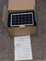 Solar panel with multiple small photovoltaic cells in black frame placed on top of black string lights inside open box with owner's manual on carpet floor.