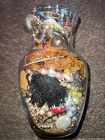 Glass jar filled with assorted jewelry showing brooches, beads, necklaces and fabric tassel