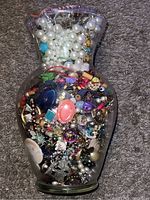 Photo of large clear glass jar filled with mixed costume jewelry including pearls, beads, brooches, and pendants.
