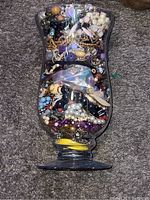 Large glass urn-shaped jar filled with assorted jewelry pieces, showing mixture of beads, chains, pendants, and rings