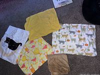 Five medium size towels displayed on the floor including a white towel with black cat, a yellow towel, a cocktail-themed towel, a patterned cat and pumpkin towel, and a brown textured towel.