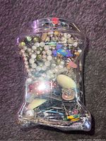 Plastic jar filled with various jewelry items including pearl necklaces, bracelets, pins, and mineral rocks viewed from one side.