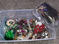 Plastic tub containing a variety of mixed jewelry items such as earrings, necklaces, bracelets, beads, and decorative rocks
