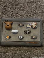 Ring case with eight rings displayed in two rows on a gray background. Front row left to right: orange/yellow gemstone ring, white band ring, eye motif ring, silver-tone striped ring. Back row left to right: reindeer head ring, silver-tone striped band ring, clear floral cluster ring, multicolored disc ring.