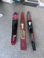 Three single water skis displayed on a garage floor: a black Vogue Ski Co. ski with bright colorful graphics, a red vintage wooden ski with textured footrest and strap, and a black Connelly SPS ski with adjustable bindings and multicolor stripes.