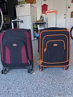 Photo shows two soft-sided carry-on suitcases side by side on a garage floor. One suitcase is black with burgundy accents, the other black with orange accents, both with telescoping handles fully extended and four spinner wheels visible.