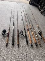 Full view of 8 fishing poles, 3 with reels attached, laid on floor for inspection showing condition and style.