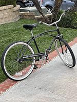 Side view of a black Huffy Good Vibrations cruiser bicycle with whitewall tires parked on a driveway next to green grass and plants.