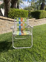 Front view of the lawn chair outdoors on grass with a tan brick wall behind. Shows multi-colored woven strips and white tubular metal frame.