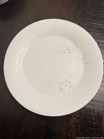 Close-up of a white plate with embossed floral rose design and silver trim on rim, showing detail of pattern and finish.