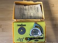 Top-down image of yellow plastic molded case holding a Powermite circular saw toy with a metal blade and instruction sheet