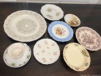Eight assorted decorative plates and one cup arranged on dark surface, showing variety of sizes, patterns and colors including Royal Doulton Tuscan Rose and Mulberry styles.