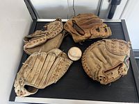 Four vintage baseball mitts and one vintage baseball displayed on a black surface, showing wear and aged leather.