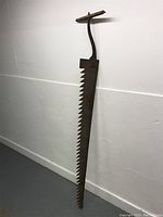 Side view of the antique ice saw showing the large jagged blade and T-shaped wooden handle.