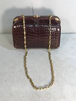 Front view of rectangular handbag showing glossy medium brown alligator-pattern leather exterior with gold-tone chain strap and round clasp closure on top.