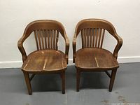 Pair of identical solid oak bankers style armchairs with curved backs, vertical slats, and contoured seats showing wear and some splits.