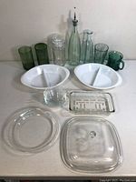 Full view of all glassware items arranged on a counter including two white divided milk glass dishes, multiple clear glass baking dishes and lids, green glass tumblers and mugs, clear glass bottles with pour spouts, a textured glass jar, and a clear decorative bowl.