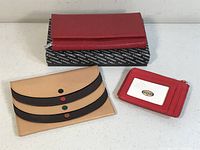 Photo showing all three wallets: a red zippered Fossil wallet, a beige and black buttoned envelope wallet, and a red card holder wallet with clear window.
