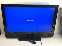 Front view of Vizio TV powered on showing 'No Signal' message on screen, remote placed on TV stand.