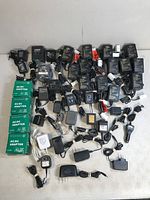 Full spread of assorted wall wart power supplies and chargers shown on surface, including several green boxed AC/DC adapters and many black adapters with cords.