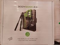 Box front showing the BodyBoss 2.0 branding and image of the gym system components including the foldable base, handles, and resistance bands.
