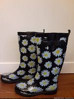 Side profile of black rubber rain boots with white daisy pattern and small black side bow on wood floor against wall.