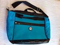 Front view of teal and black Foxy Jeans bag showing adjustable shoulder strap, top handle, front zippered pocket, and overall design.