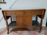 Front view of the Gibbard solid wood server/bar cart showing the marble top insert, two drawers, drop-down sides folded down, double-door cabinet and lower shelf.