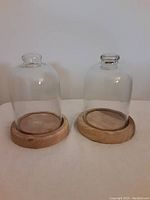 Pair of glass cloches with wooden bases shown side by side on a flat surface against a plain wall background.