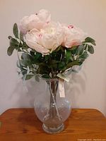 Faux peony bouquet in a clear glass vase on wooden surface against plain background