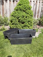 Set of three black plastic rectangular deck railing planter boxes shown on grass with wood fence and greenery in background.