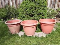 Three large reddish-orange plastic flower pots placed on grass with a pine bush background, showing front exterior sides.