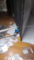 Photo of a pear-shaped cabochon black opal gemstone showing vivid blue and green flashes held by tweezers in natural light.