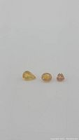 Three natural sapphires in yellow and orange colors, pear, oval, and heart cuts lined up on a white background.