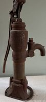 Front view of antique cast iron hand water pump showing overall shape, lever handle, spout, and rust condition.