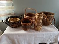 Six assorted woven baskets displayed on a white tablecloth, varying in size, shape and color, highlighting the variety of weave styles and handle types.