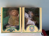 Two Cabbage Patch Kids dolls in original boxes, one in a yellow dress and one in a white Preemie outfit.