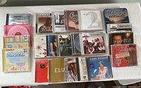 Photo shows 22 assorted music CDs laid out on a white surface, including genres like jazz, country, rock, pop, compilations, and baby lullabies.