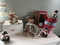 Wide view of Christmas lot on white table including Santa music box, cookie tins, snowmen and gingerbread bake set box