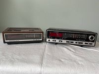 Two vintage clock radios, one with wood grain casing and the other a Sears Comtrek II model, both placed side by side on a white cloth background.
