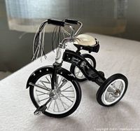 Black decorative tricycle model with white seat, chrome pedals, and black and white handlebar streamers on white surface.