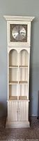 Full view of tall white grandmother clock style cabinet with decorative clock face, shelves, and doors