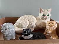 Four ceramic cat figurines arranged in a cardboard box, showing size differences and color details.