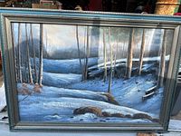 Photo of a large framed painting showing a winter snow-covered forest landscape with leafless trees and ground covered with snow and some fallen logs.