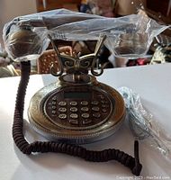Front and top view of an elegant vintage-style telephone showing ornate metal design, plastic-wrapped handset, and push button keypad.