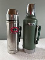 Photo shows two used metal thermos bottles: one stainless steel with a red Unovad logo, one green Stanley thermos with a metal cup lid and handle.