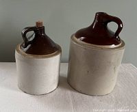 Pair of two vintage ceramic jugs showing brown glazed top with handle and lighter unglazed base, placed side by side.