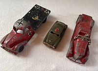 Photo shows three vintage toy vehicles: red German military truck with flatbed, green US Army car, and red classic car with significant paint chipping.