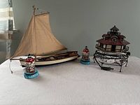 Wooden sailboat with fabric sail placed next to one large lighthouse with electrical cord and two smaller lighthouse figurines on a white cloth.